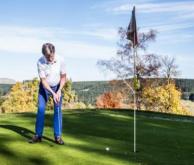 Golfer beim Abschlag, Ball liegt kurz vor dem Zielloch, im Hintergrund schöne Herbstlandschaft